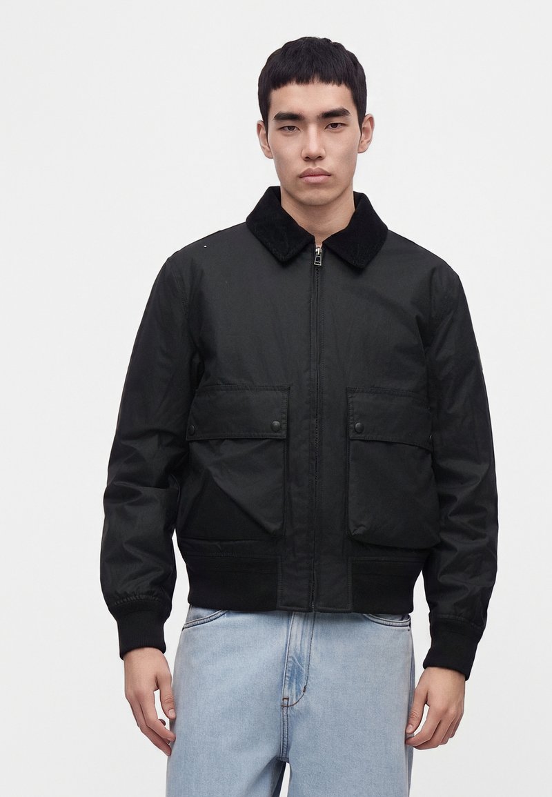Jeune homme portant une veste noire zippée avec un col et deux poches avant, associée à un jean bleu clair, debout devant un fond uni.