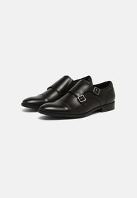 Zapatos de cuero negro con punta redondeada, doble hebilla ajustable, textura suave y un tacón bajo apilado, presentando un diseño minimalista.