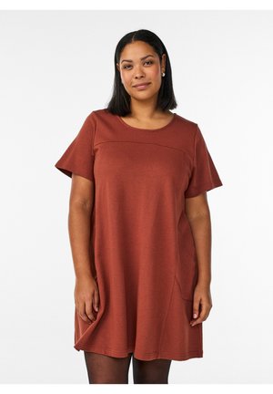 Kvinde med mellem hudtone og lige sort hår iført en rustfarvet, kortærmet kjole stående mod en hvid baggrund.