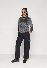 Szara bluza polarowa na suwak z wysokim kołnierzem, w zestawie z czarnymi spodniami cargo z wieloma kieszeniami. Akcesoria to białe okulary przeciwsłoneczne i czarne buty.