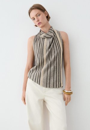 Vrouw draagt mouwloze beige en grijze top met verticale strepen, gedrapeerde kraag en ecru wijde broek, poserend tegen een witte muur.