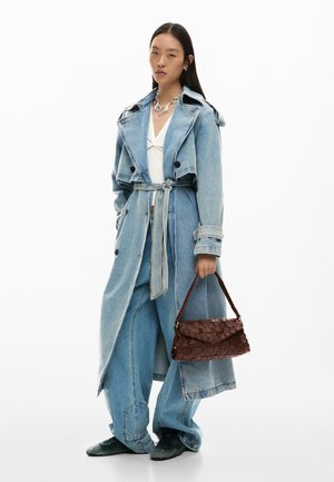 Jauna moteris dėvi šviesiai mėlyną džinsinį tranšėjos paltą ir plačias džinsines kelnes, rankoje laiko rudą pintą rankinę, stovi ant balto fono.