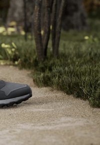 Black athletic shoe stepping on a sandy outdoor trail with green grass and tree trunks in the background.
