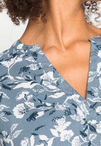 Blusa con stampa floreale caratterizzata da uno sfondo grigio con disegni bianchi e blu scuro, scollatura a V e tessuto morbido.