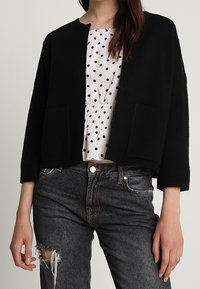 Kvinna i en svart cardigan med öppen front över en vit blus med prickar och slitna mörka jeans mot en enkel bakgrund.