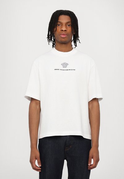 Tricou alb cu mânecă scurtă, realizat din bumbac, având un print grafic cu un logo Medusa și textul "VERSACE" dedesubt. Gât clasic de tip crew.