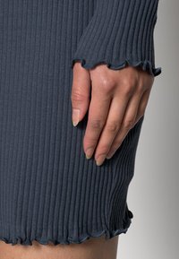 Ribbad stickad klänning i mörkgrå, med en vågig kant vid ärmarna och nederkanten. Hand placerad på sidan av klänningen.