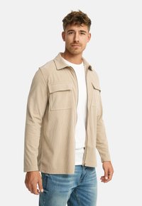 Mand iført en beige fløjljakke over en hvid skjorte og blå jeans, stående mod en hvid baggrund.