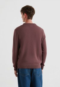 Maglione lavorato a maglia marrone con colletto e polsini a costine, abbinato a jeans a gamba larga blu, visto da dietro. La texture appare morbida e calda.