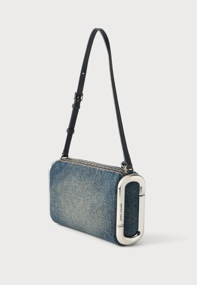 Sac à bandoulière rectangulaire en denim bleu avec une sangle noire réglable et un grand détail ovale en métal argenté gravé Marc Jacobs.