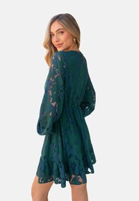 Abito in pizzo di un verde intenso, con maniche lunghe, vita aderente e gonna a campana. Motivo floreale testurizzato con orlo arricciato.