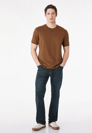Ung mand stående med hænderne i lommerne iført brun t-shirt, mørkeblå jeans og hvide sneakers mod en hvid baggrund.