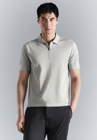 Homme portant un polo à manches courtes gris clair avec une fermeture éclair à l'avant, associé à un pantalon noir, debout devant un fond uni gris clair.