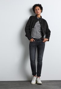 Svart mockajacka med knappstängning, randig t-shirt, mörka jeans och vita sneakers. Upprullade ärmar på jeansen, stående pose mot en vägg.