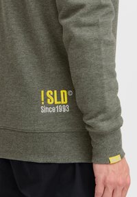 Olive green sweatshirt made of soft fabric. Features yellow and white logo detailing with "Since 1993" text on the lower left side.