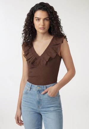 Donna con capelli ricci che indossa un top marrone senza maniche con scollo a V arricciato e jeans a vita alta azzurri, mano in tasca, espressione neutra.