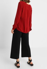 Blusa larga de manga larga texturizada en rojo, combinada con pantalones negros de pierna ancha. Sandalias negras con tiras alrededor del tobillo completan el conjunto.