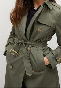 Femme portant un trench-coat ceinturé vert olive avec des boutons en écaille de tortue et des détails de boucle, les mains dans les poches du manteau.