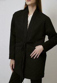 Cappotto lungo nero con colletto a scialle, vita con cravatta e due tasche frontali. Realizzato in tessuto morbido con una texture liscia, presenta una silhouette leggermente strutturata.
