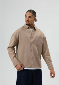 Homme aux cheveux tressés portant une chemise beige boutonnée par-dessus un t-shirt blanc et un pantalon foncé, regardant vers sa gauche sur un fond uni.