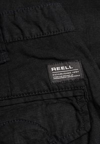 Schwarzer Stoff mit einem subtilen Gittermuster, versehen mit einem Etikett, das "REELL ESTABLISHED 1997" in weißer Schrift auf schwarzem Hintergrund zeigt.