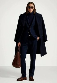 Temno modri volneni plašč prek ujemajočega se obleka, kombiniran s temnim puloverjem z ovratnikom. Dodatki vključujejo sončna očala in rjavo usnjeno torbo. Rjavi čevlji.