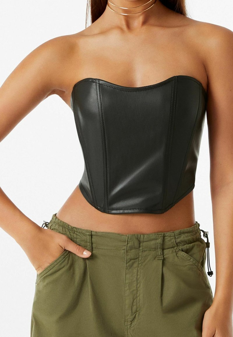 Femme portant un corset en cuir noir sans bretelles et un pantalon vert olive, les mains dans les poches, sur un fond uni.