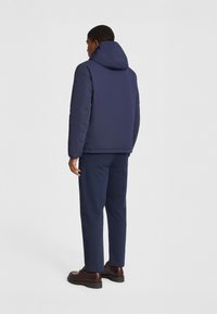 Giubbotto puffer blu navy con cappuccio, caratterizzato da una texture liscia e una toppa con logo sulla spalla sinistra. Abbinato a pantaloni blu navy e stivali marroni.
