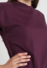 Bluza z krótkim rękawem w głębokim purpurowym kolorze, wykonana z bawełny, z okrągłym dekoltem i luźnym krojem, o gładkiej fakturze. Widać rękę opartą na biodrze.