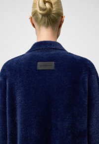 Navyblauer Kunstpelzmantel mit Schalkragen, mit einem schwarzen Etikett auf der Rückseite, weicher Textur und einem leicht übergroßen Design.