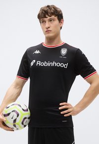 Maillot de football noir avec des accents rouges, comportant un logo et un texte sponsorisé. Le maillot a des manches courtes et un col rond, fabriqué en tissu texturé.