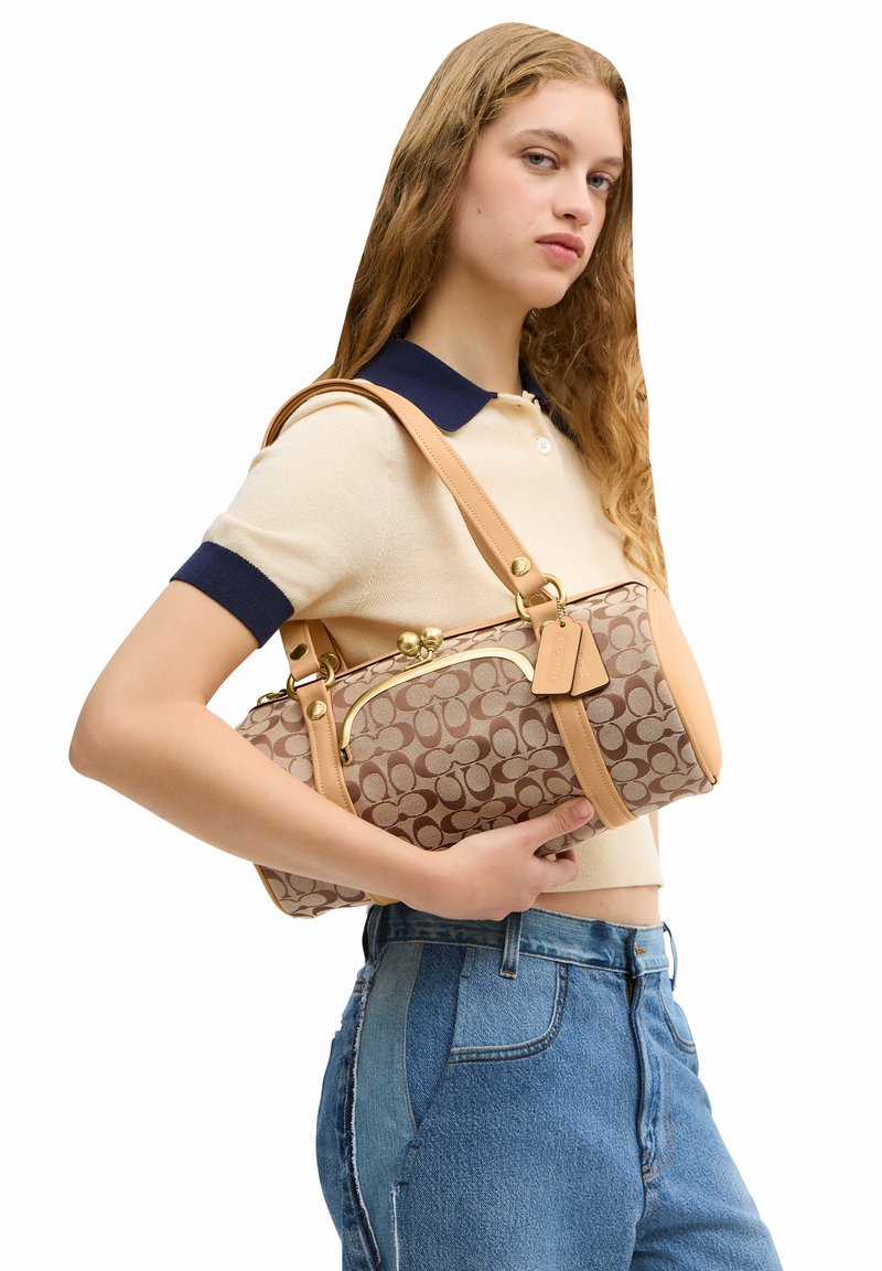 Jeune femme aux longs cheveux porte un polo beige et un jean bleu, tenant un sac à main marron à motifs sur son épaule devant un fond blanc.