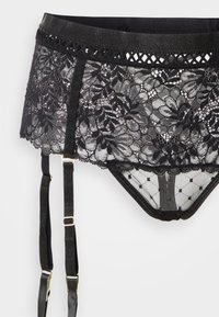 Jarretière en dentelle noire avec motif floral, présentant un tulle transparent et des pois. Comprend des bretelles réglables et des accents métalliques.