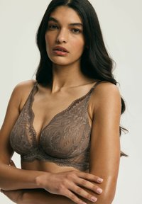 Mladá žena s dlhými tmavými vlasmi, oblečená v hnedom čipkovanom podprsenke, stoji s prekríženými rukami pred jednoduchým pozadím.