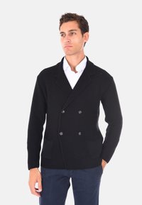 Cardigan nero a maglia a doppio petto con una finitura testurizzata, caratterizzato da due tasche frontali e bottoni neri, indossato sopra una camicia bianca.