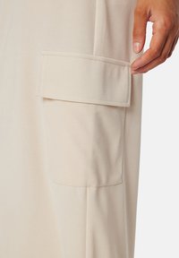 Beige stof met een gladde textuur, voorzien van een klepzak en subtiele stiksels. Het ontwerp benadrukt functionaliteit en eenvoud.
