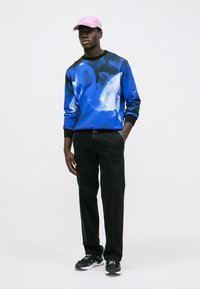 Hombre con una gorra rosa, sudadera de patrones azules, pantalones negros con costuras visibles y deportivas negras, de pie contra un fondo blanco liso.