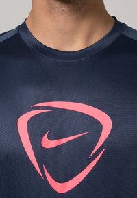 Tummansininen urheilupaita, jossa on pyöreä pääntie, keskiössä suuri vaaleanpunainen Nike-logo ja lyhyet hihat. Sileä pinta.