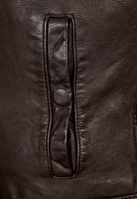 Tumma ruskea nahka, jossa on ryppyinen pinta, sisältää pystysuoran läppätaskun, joka on suljettu nepparilla ja korostaa yksityiskohtaista ompelua.