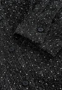Svart texturerad tygskjorta med ett vävt mönster, som har små vita prickar. Inkluderar svarta knappar vid ärmarnas och framfickans öppningar.