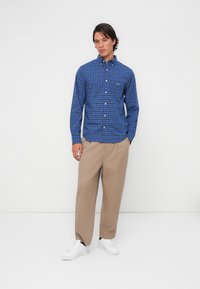 Blue checked shirt with long sleeves, button-up collar, and chest pocket. Worn with beige wide-leg trousers and white sneakers.