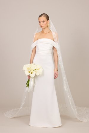 Mariée en robe de mariage blanche épaules dénudées avec long voile en dentelle tenant un bouquet de callas blancs sur un fond simple.