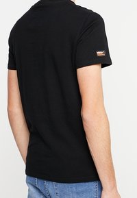 Camiseta negra de manga corta hecha de algodón, con cuello redondo y un pequeño parche de logo en la manga izquierda. Corte clásico y textura suave.