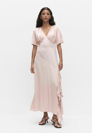 Nainen seisoo päällään vaaleanpunainen satiininen kietaisumekko, jossa lyhyet hihat, röyhelödetaalit sekä mustat remmikkäät korkokengät.