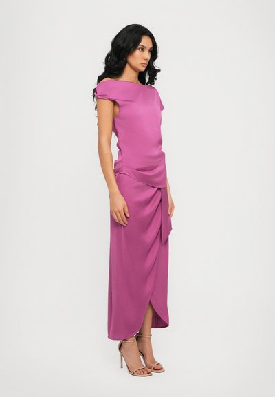 Robe fuchsia avec un design drapé, manches courtes et une fente latérale. Fabriquée en tissu lisse, associée à des talons à bride de cheville couleur beige.
