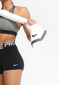 Shorts de sport noirs avec logo Nike blanc, brassière de sport grise, et serviette blanche avec accent gris. Serviette tenue dans une main.