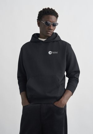 Mees, kes kannab musta New Balance kapuutsiga pusa ja tumedaid pükse, käed taskutes, väikeste ovaalsete päikeseprillidega, seisab heleda tausta ees.