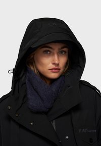 Zwarte waterdichte jas met een grote capuchon, gouden hardware accenten, en een donkerblauwe gebreide sjaal om de nek.