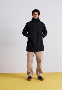 Zwarte waterdichte jas met hoge kraag, ritssluiting en zijzakken, gecombineerd met beige broek en zwarte schoenen op een gele vloer.