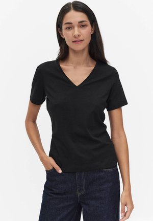 Vrouw met lang donker haar die een zwart V-hals shirt met korte mouwen en donkerblauwe spijkerbroek draagt, staand met één hand in haar zak.